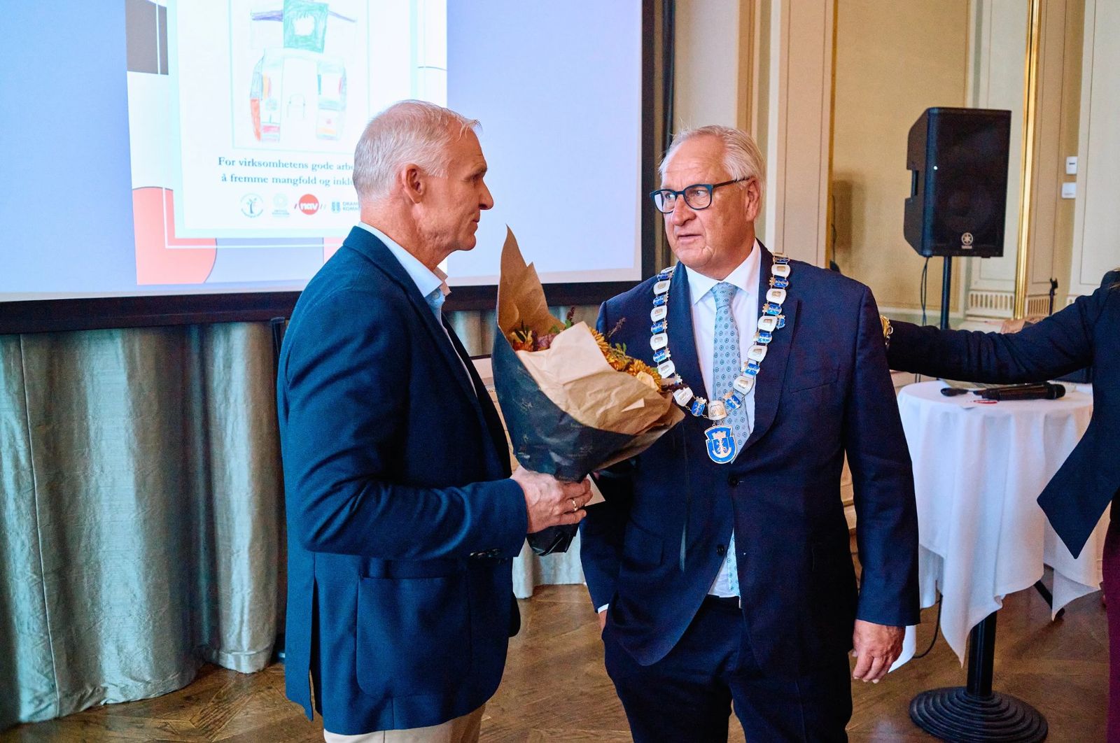 Ordføreren, iført kjede, overrekker blomster til representanten for Hitachi Energy etter at bedriften vant Mangfoldsprisen 2026.